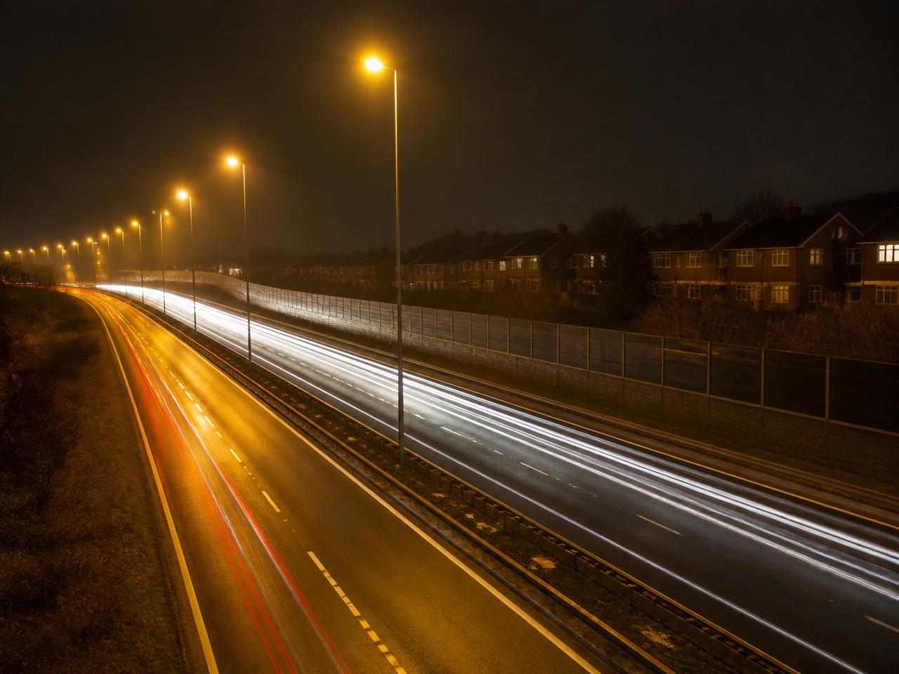 New dual carriageway at night with tall street lighting and traffic light trails beside a housing estate